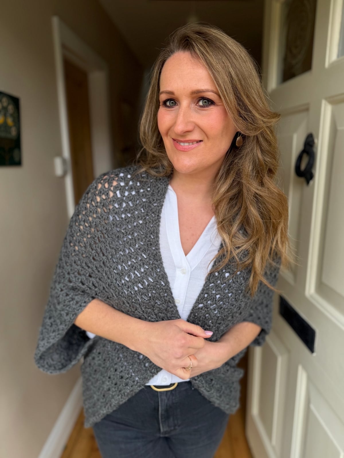 A woman with long, wavy blonde hair stands indoors by a white door, wearing a grey knitted shawl over a white blouse and dark pants, smiling at the camera.