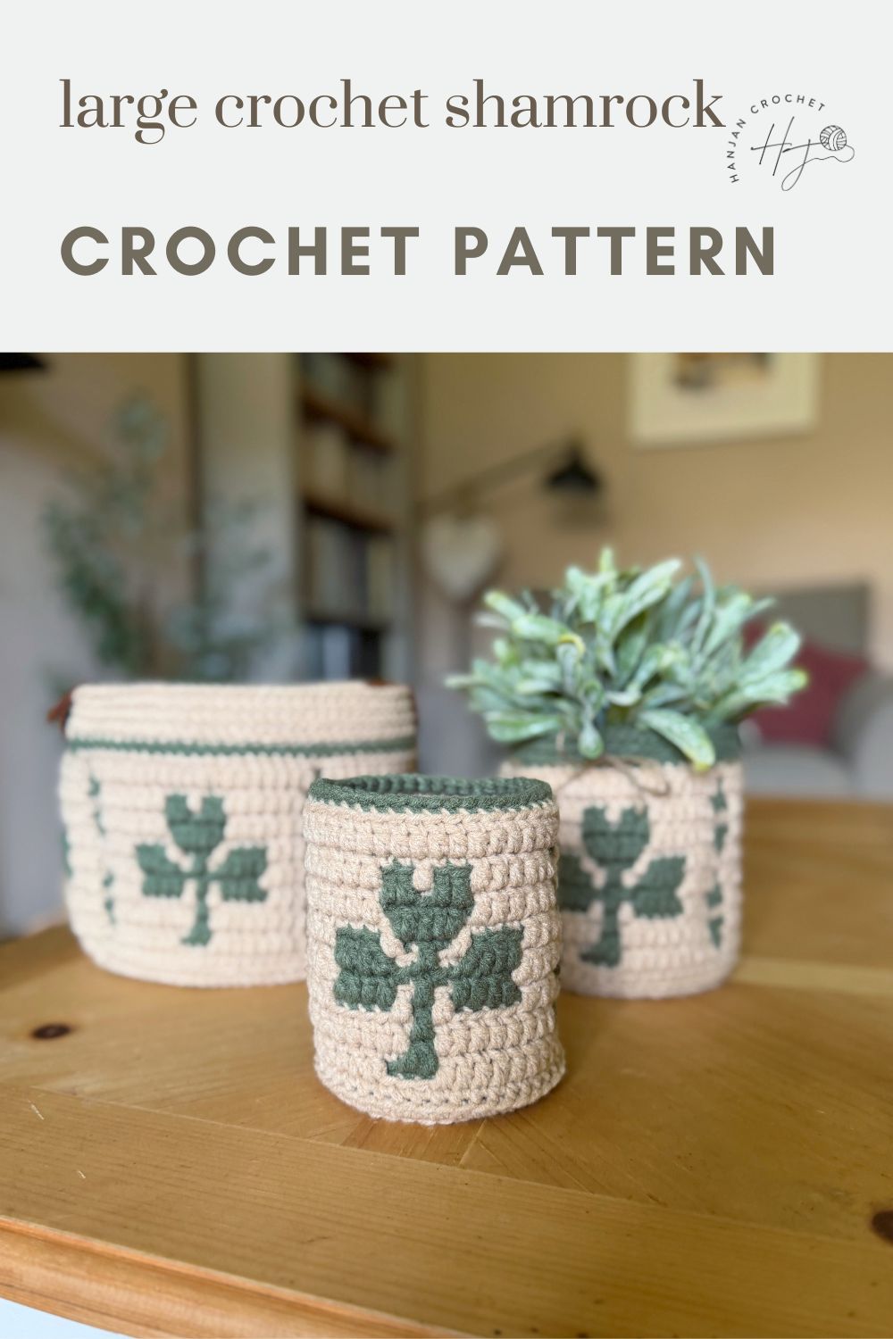 Three beige crochet containers with green shamrock designs are displayed on a wooden table, with a plant in the background. Text reads: "large crochet shamrock CROCHET PATTERN.