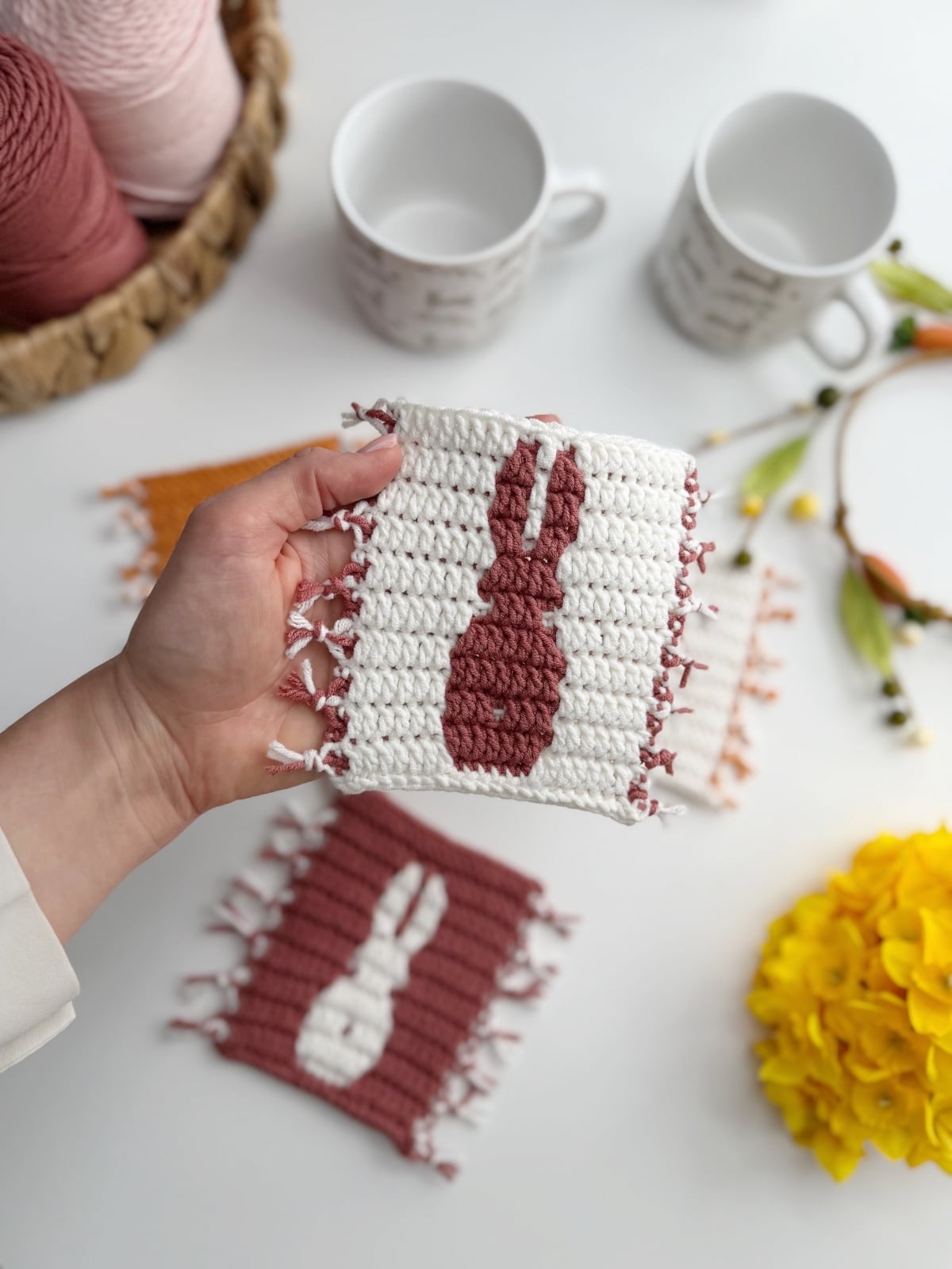 A hand holds a crocheted coaster with a pink bunny silhouette; more crocheted coasters, mugs, and flowers are on the white table in the background.