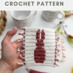 A hand holds a rectangular crochet coaster featuring a rabbit silhouette design, made using a charming crochet bunny coaster pattern, with mugs and more coasters in the blurred background.