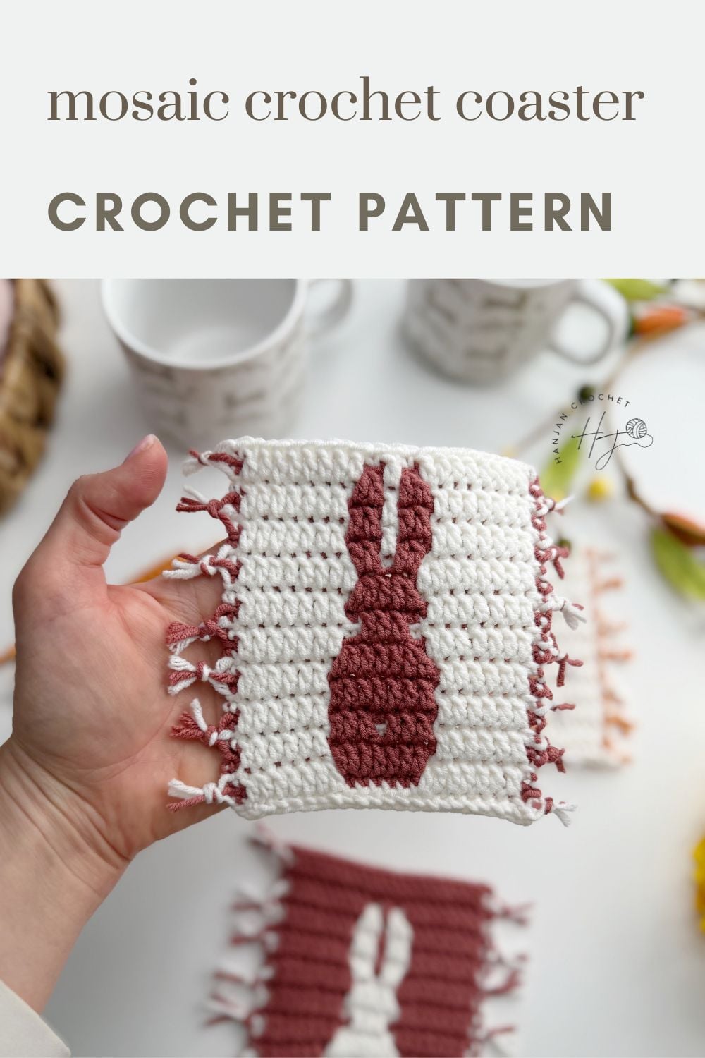 A hand holds a rectangular crochet coaster featuring a rabbit silhouette design, made using a charming crochet bunny coaster pattern, with mugs and more coasters in the blurred background.