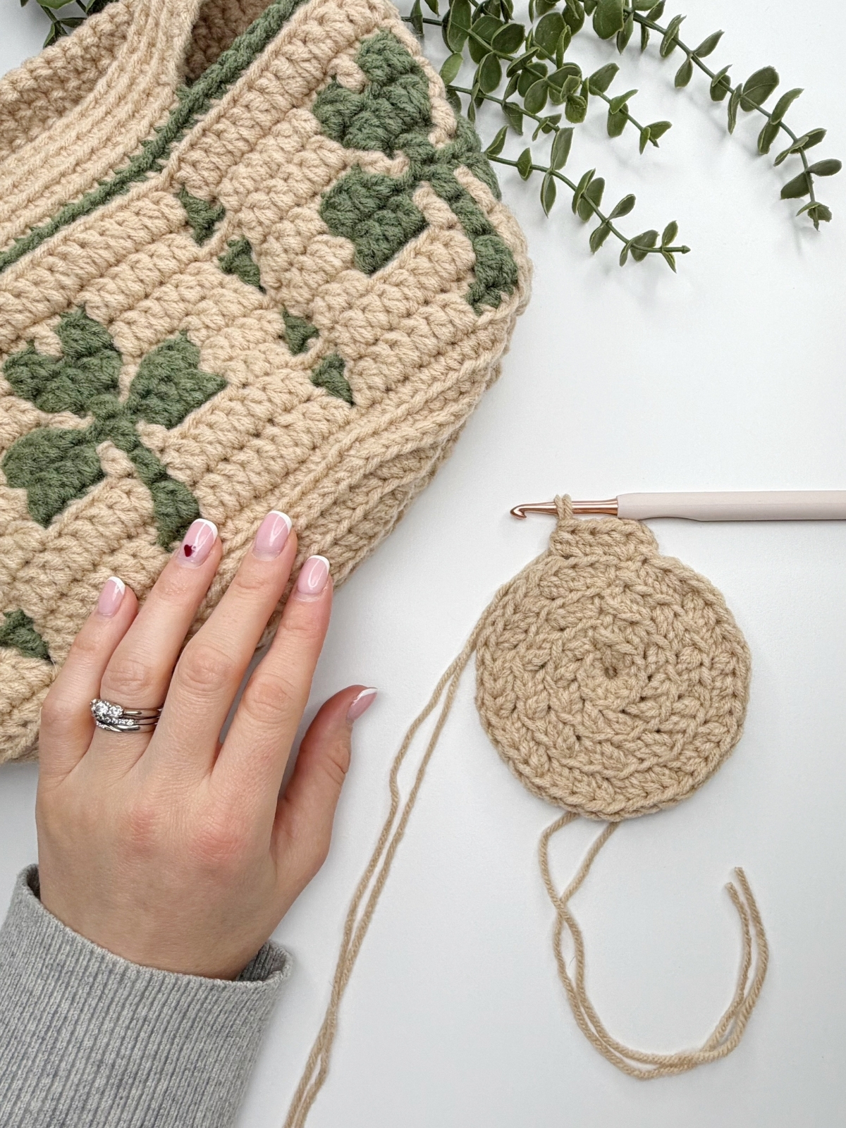 A hand with a ring rests on a crocheted bag with green and beige pattern next to a crocheted shamrock, a beige crochet circle, and a crochet hook on a white surface with greenery in the background.