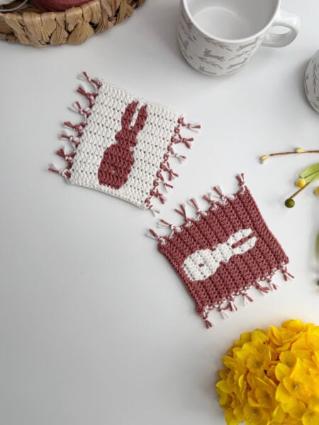 Two rectangular crocheted coasters with bunny silhouettes, one in white with a pink bunny and one in pink with a white bunny, placed on a white surface near mugs and yellow flowers.