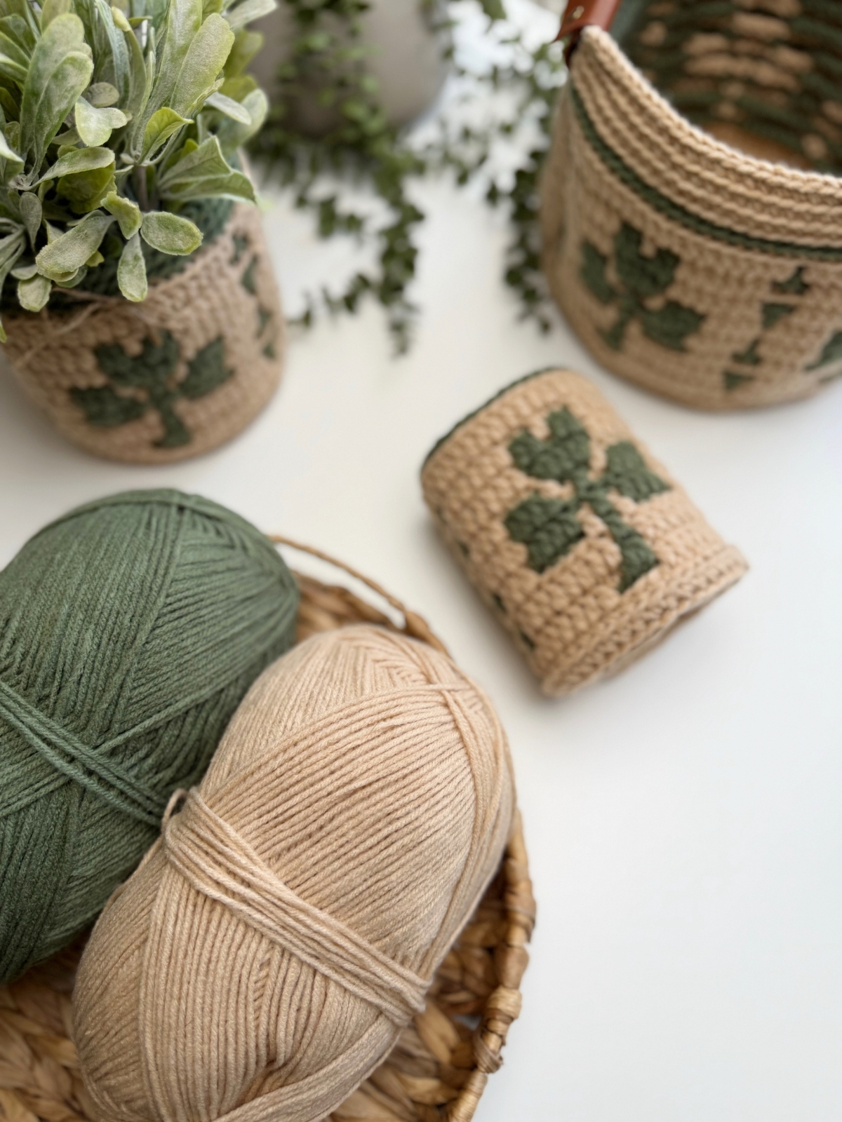 Two skeins of yarn, one green and one beige, sit in a woven basket next to crocheted containers with green leaf patterns, a crocheted shamrock, and potted plants on a white surface.