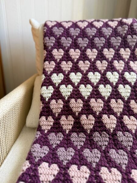 A crocheted blanket with rows of heart patterns in shades of purple, pink, and cream—made using a free crochet heart blanket pattern—is draped over a beige armchair.