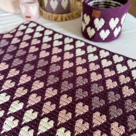 Crocheted blanket and baskets in shades of purple, pink, and cream, all featuring heart patterns, displayed on a bed&mdash;made using a free crochet heart blanket pattern.