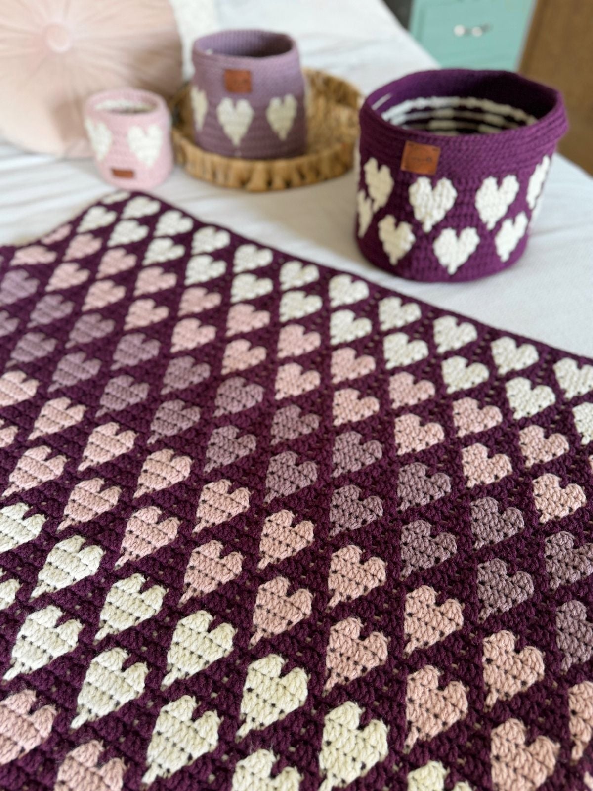 Crocheted blanket and baskets in shades of purple, pink, and cream, all featuring heart patterns, displayed on a bed—made using a free crochet heart blanket pattern.