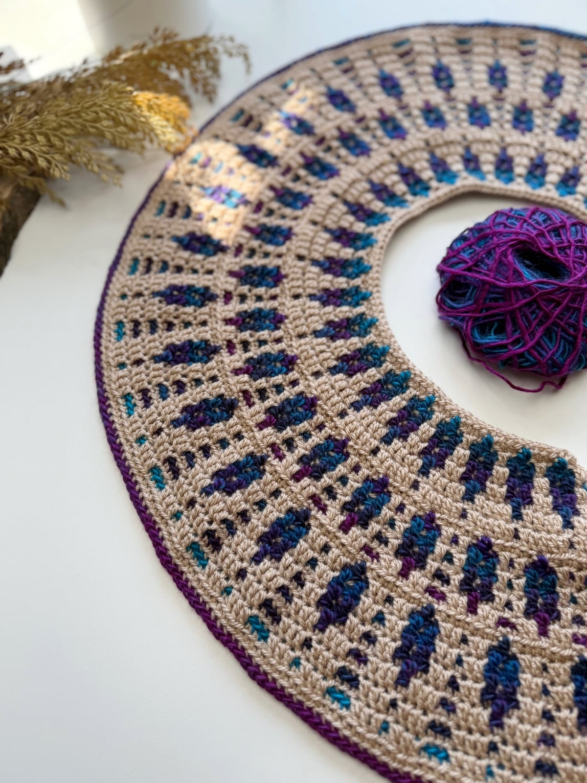 A round crochet project with a beige base and blue, purple, and teal accents&mdash;reminiscent of a mosaic crochet cardigan pattern&mdash;sits beside a matching yarn ball on a white surface with dried plants in the background.