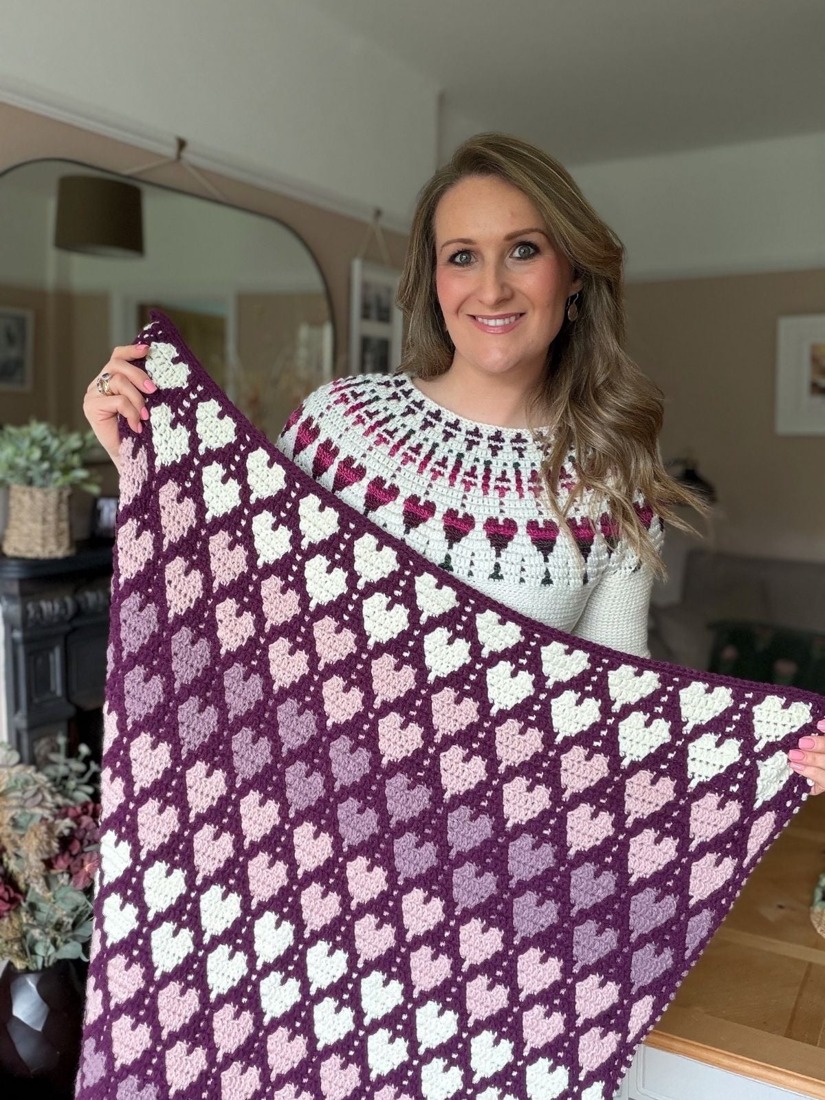 A woman stands indoors, smiling and holding a crocheted blanket with a purple and pink geometric design inspired by a free crochet heart blanket pattern.