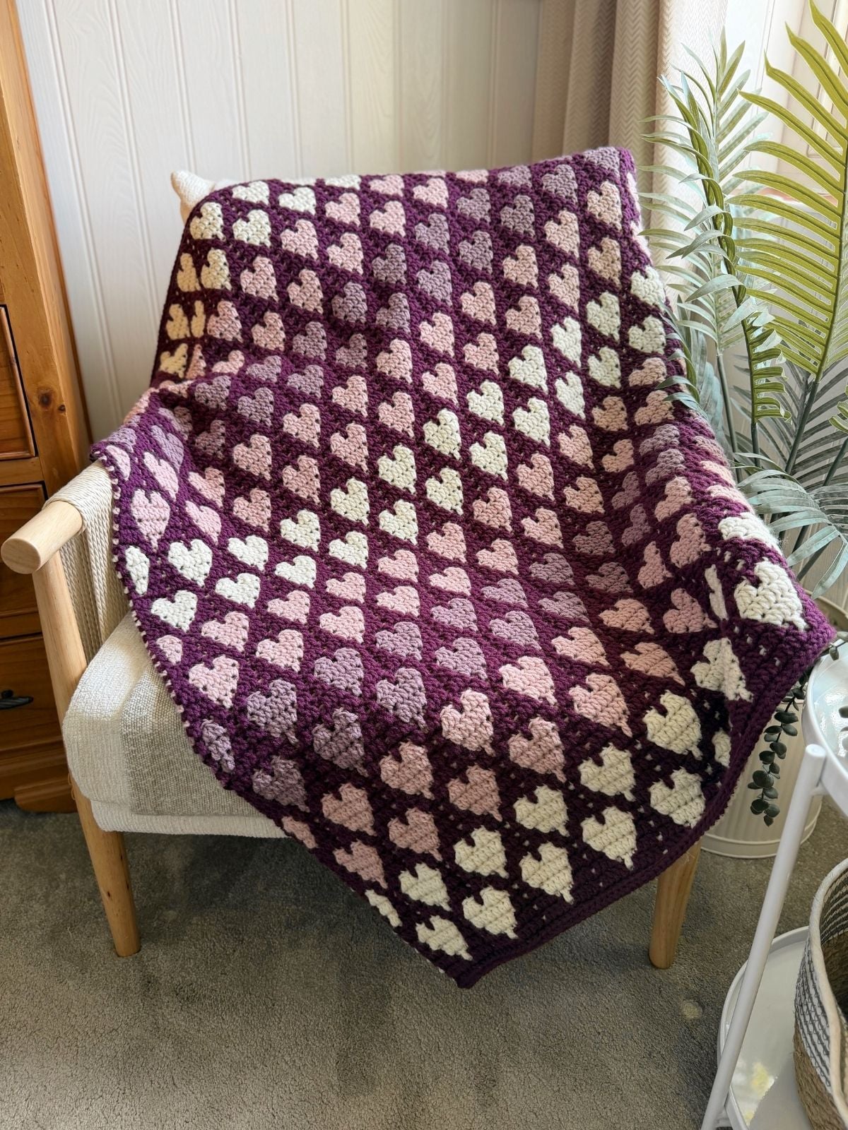 A crocheted blanket with a repeating heart pattern in shades of purple, pink, and cream draped over a wooden armchair.