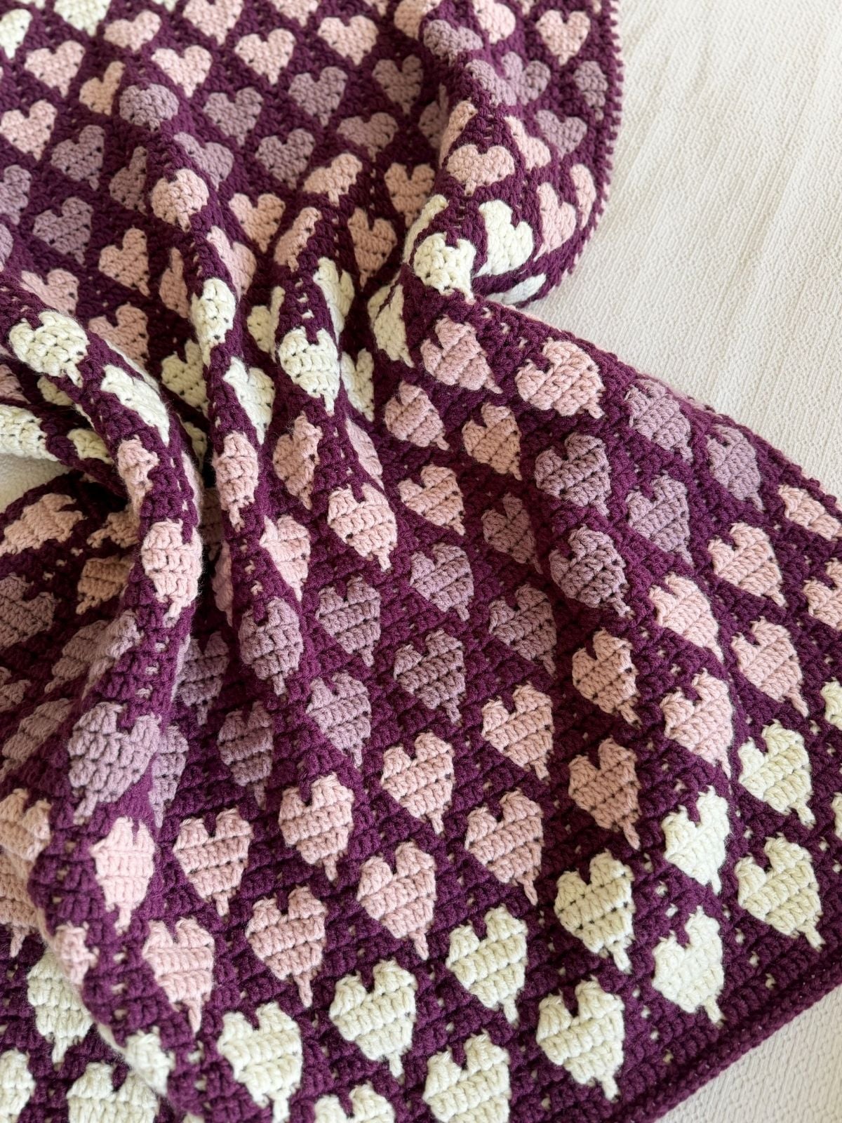 A crocheted blanket featuring a repeating pattern of small heart shapes in shades of purple, pink, and cream, draped over a light-colored surface—perfect inspiration if you're searching for a free crochet heart blanket pattern.