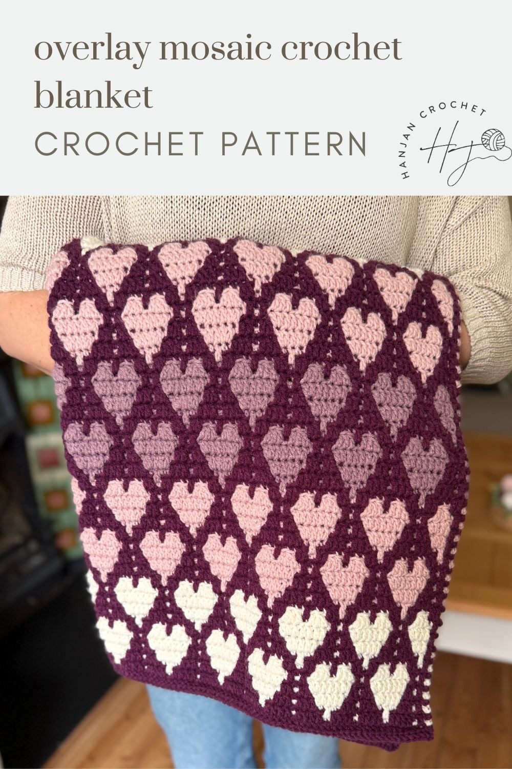 Person holding a crochet blanket featuring a repeating heart pattern in shades of purple, pink, and cream. Text reads “overlay mosaic crochet blanket crochet pattern.”.