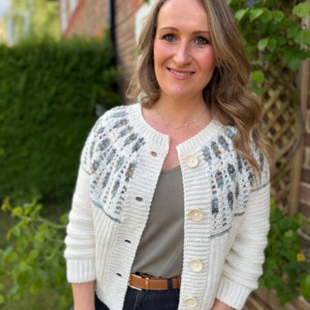 A woman with wavy blonde hair stands outside near a brick house and greenery, wearing a patterned white mosaic crochet cardigan, gray shirt, and black pants.