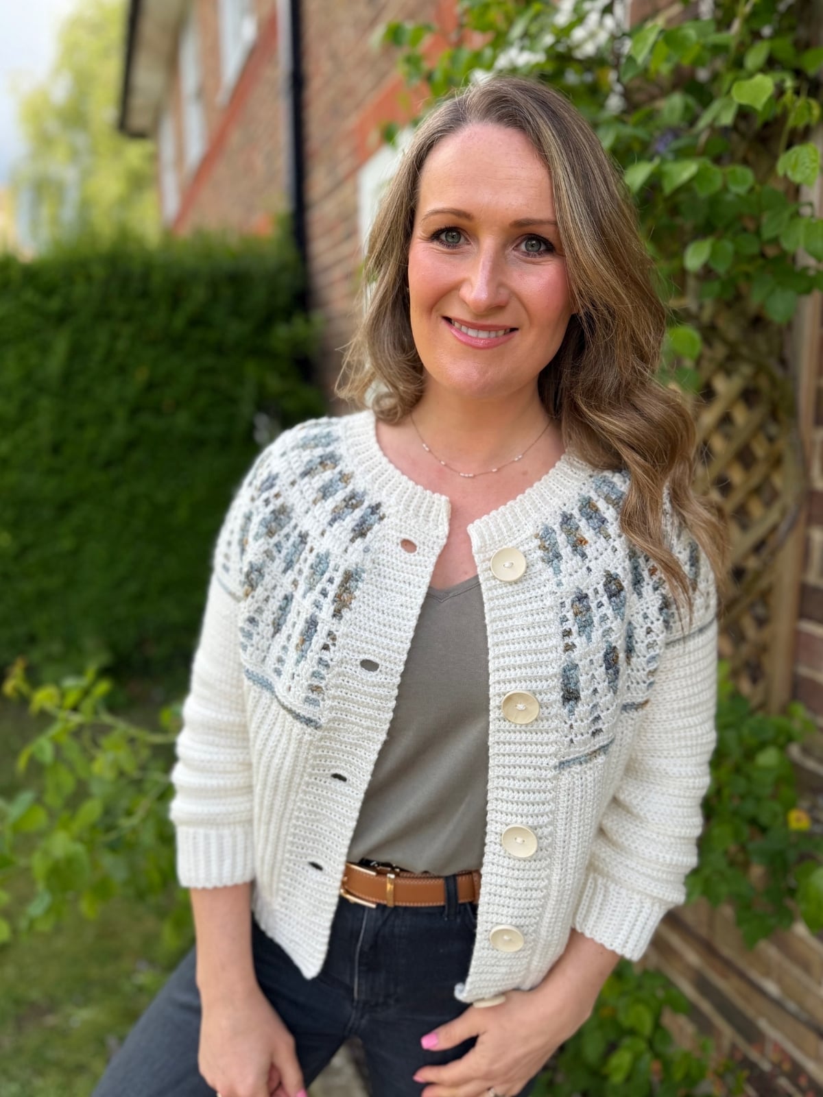 A woman wearing a cream mosaic crochet cardigan with patterned details stands outside near a brick house, smiling at the camera.