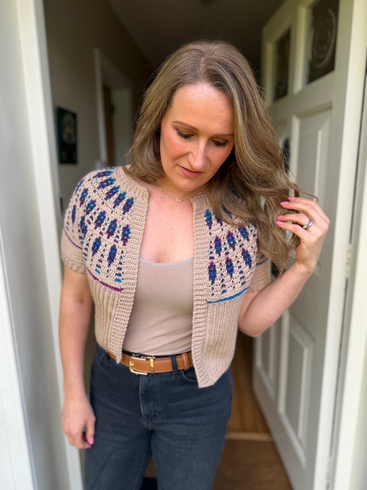 A woman stands indoors near a doorway, wearing a beige short-sleeve knit cardigan featuring a mosaic crochet cardigan pattern, paired with a tan top and dark jeans.