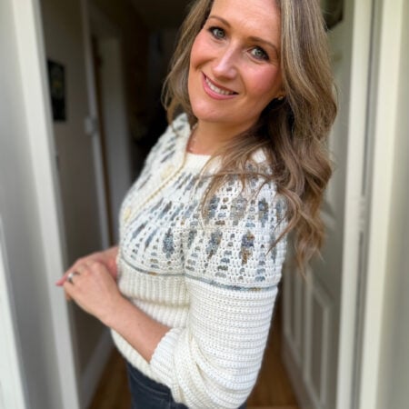 A woman with long, wavy hair stands indoors, smiling at the camera. She wears a white and gray mosaic crochet cardigan pattern sweater and dark pants.
