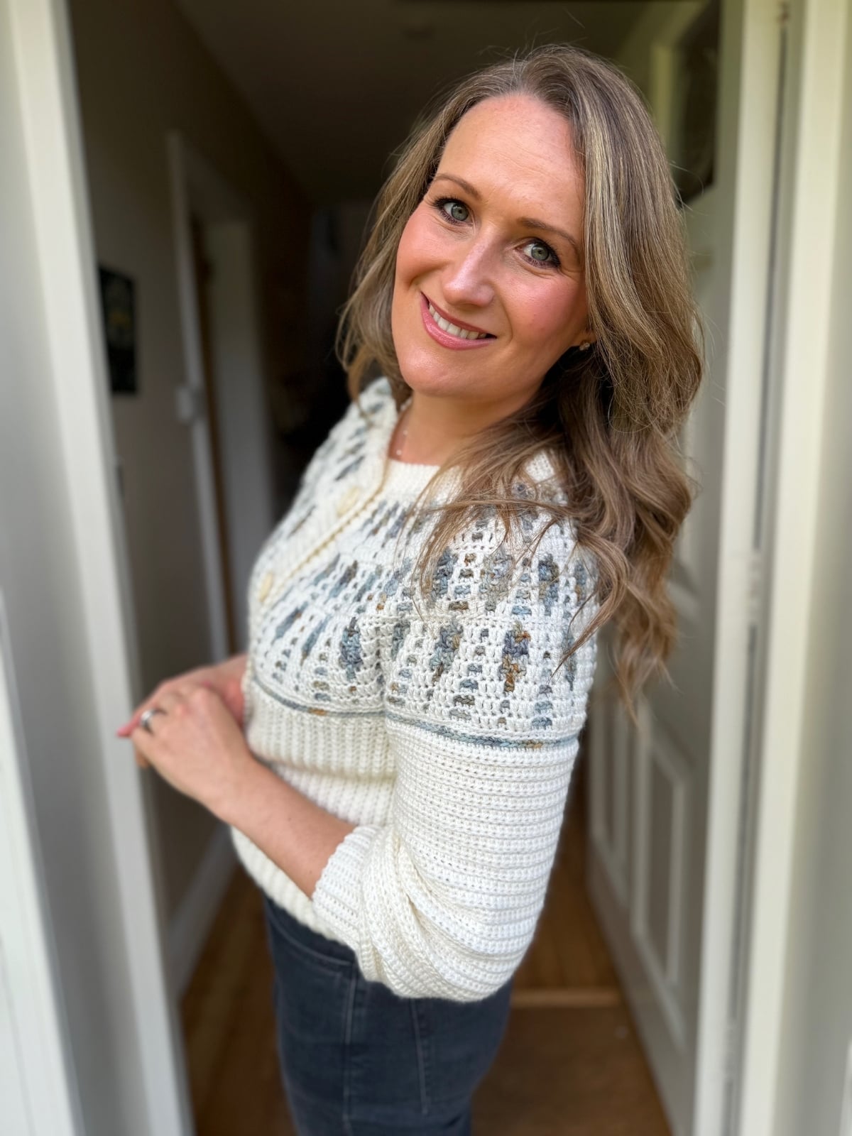 A woman with long, wavy hair stands indoors, smiling at the camera. She wears a white and gray mosaic crochet cardigan pattern sweater and dark pants.
