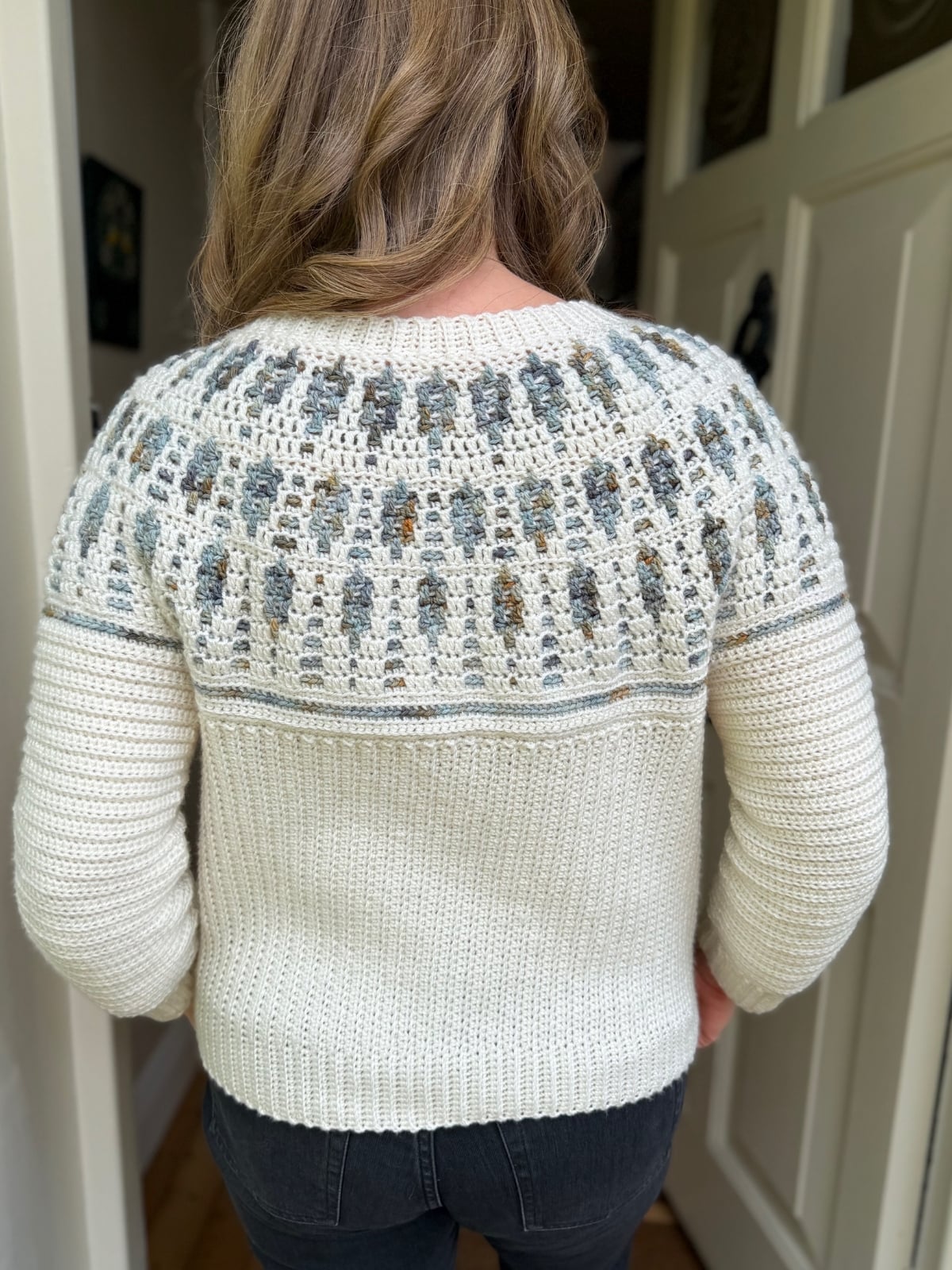 A person stands indoors wearing a cream knit sweater with a patterned yoke in gray and blue tones, reminiscent of a mosaic crochet cardigan pattern, viewed from the back.