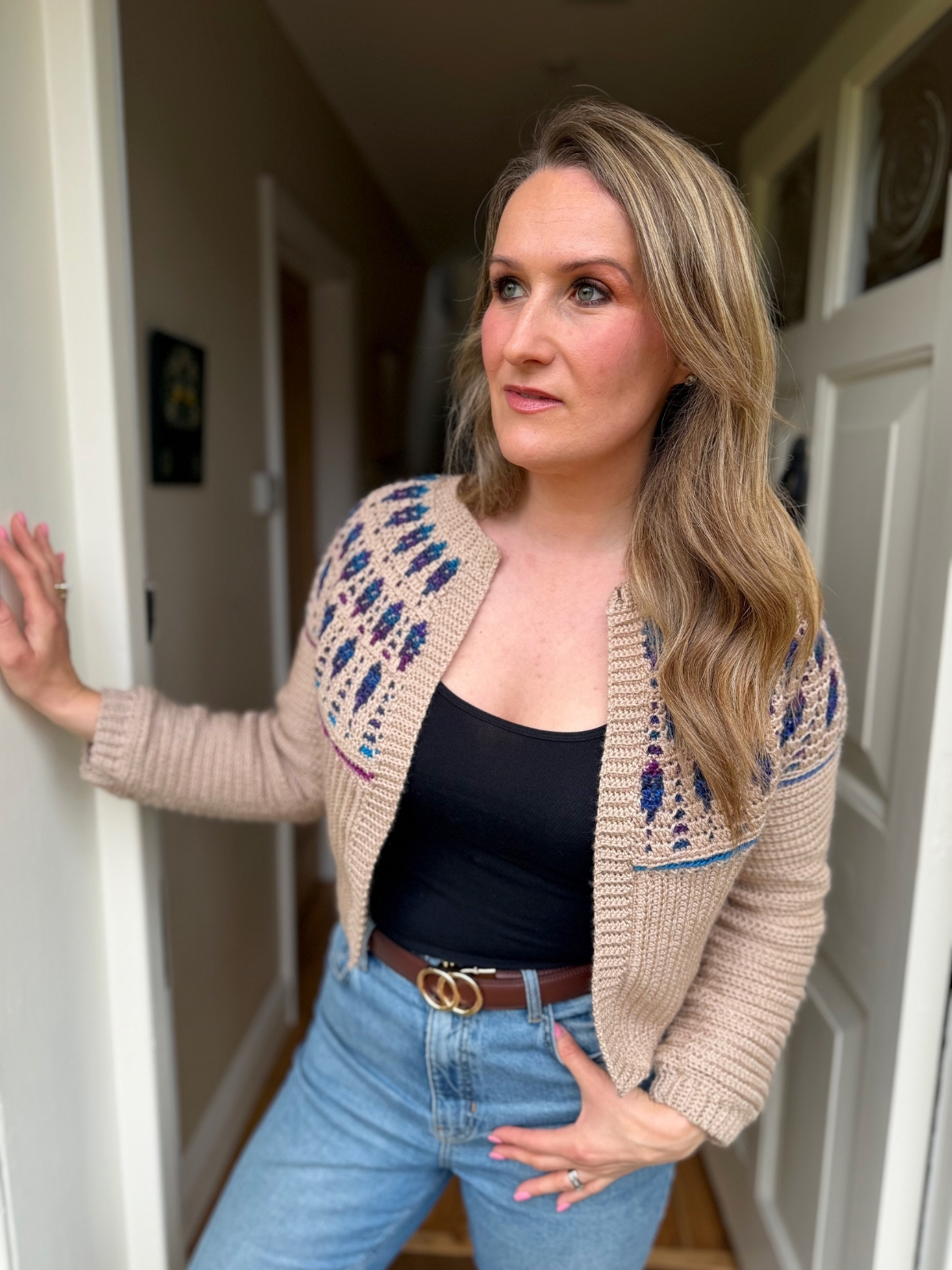 A woman with long blonde hair stands in a hallway, wearing a mosaic crochet cardigan pattern over a black top and blue jeans, with one hand resting on a door frame.