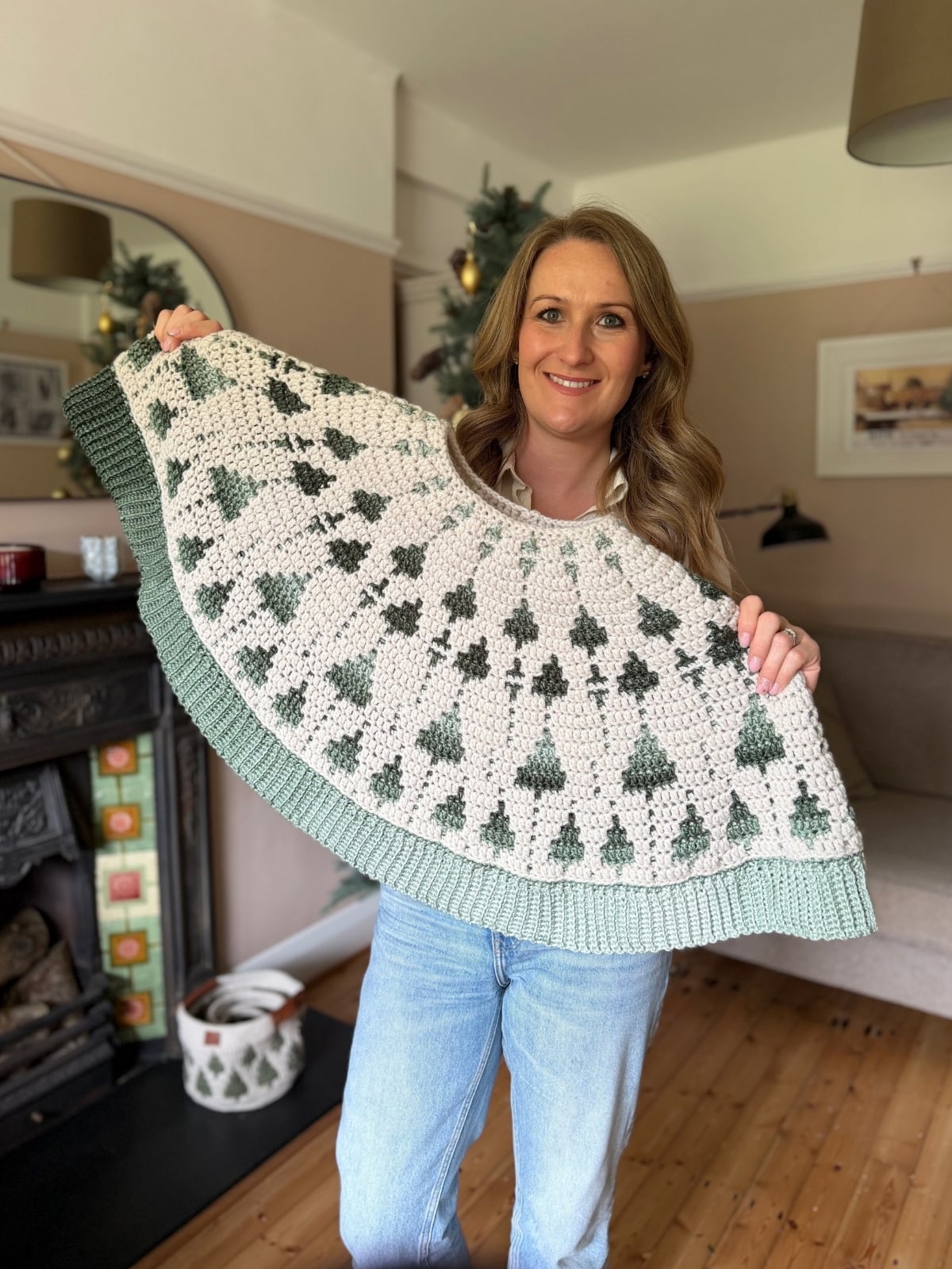 A woman stands in a living room holding up a crochet, circular tree-patterned garment with white and green colors, resembling a beautiful crochet tree skirt pattern.