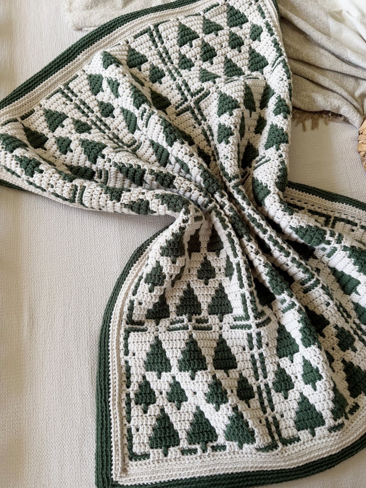 A crocheted blanket with green and cream geometric and tree patterns is laid out on a light-colored surface.