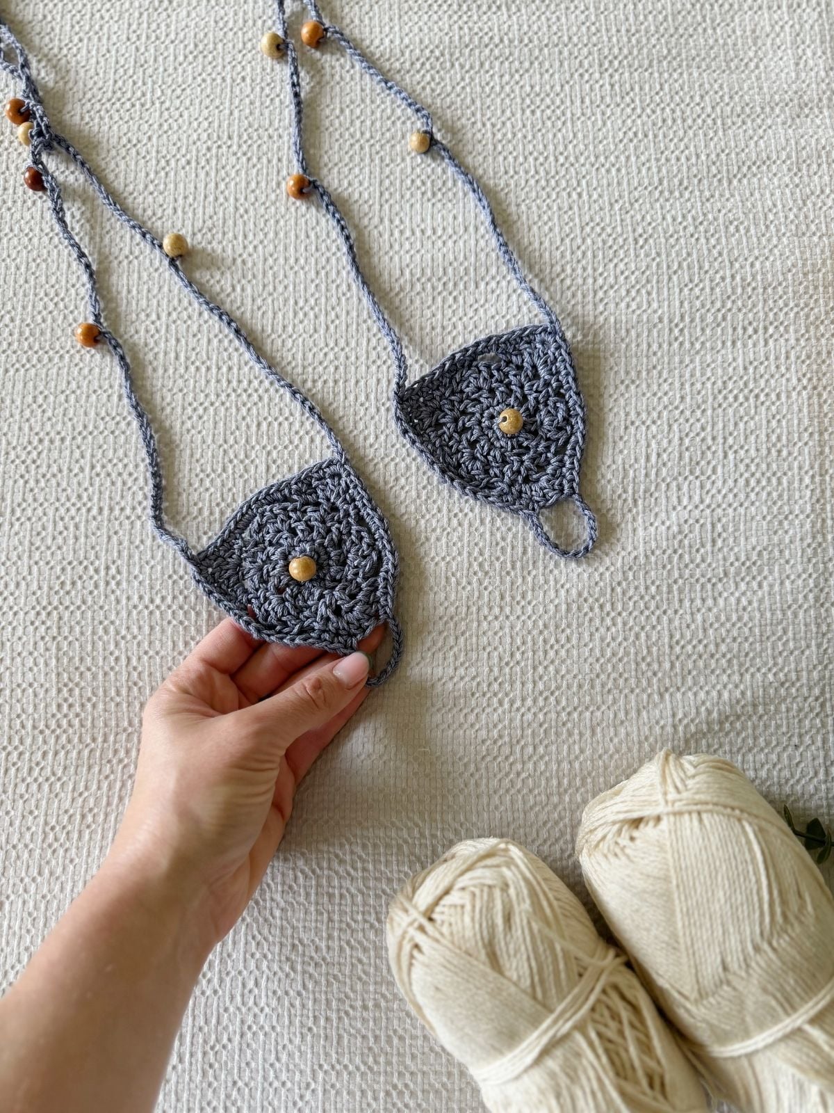 A hand holds a crocheted gray face mask with beads, next to another mask and two skeins of cream yarn on a textured surface—perfect materials for trying out a free barefoot crochet sandals pattern.