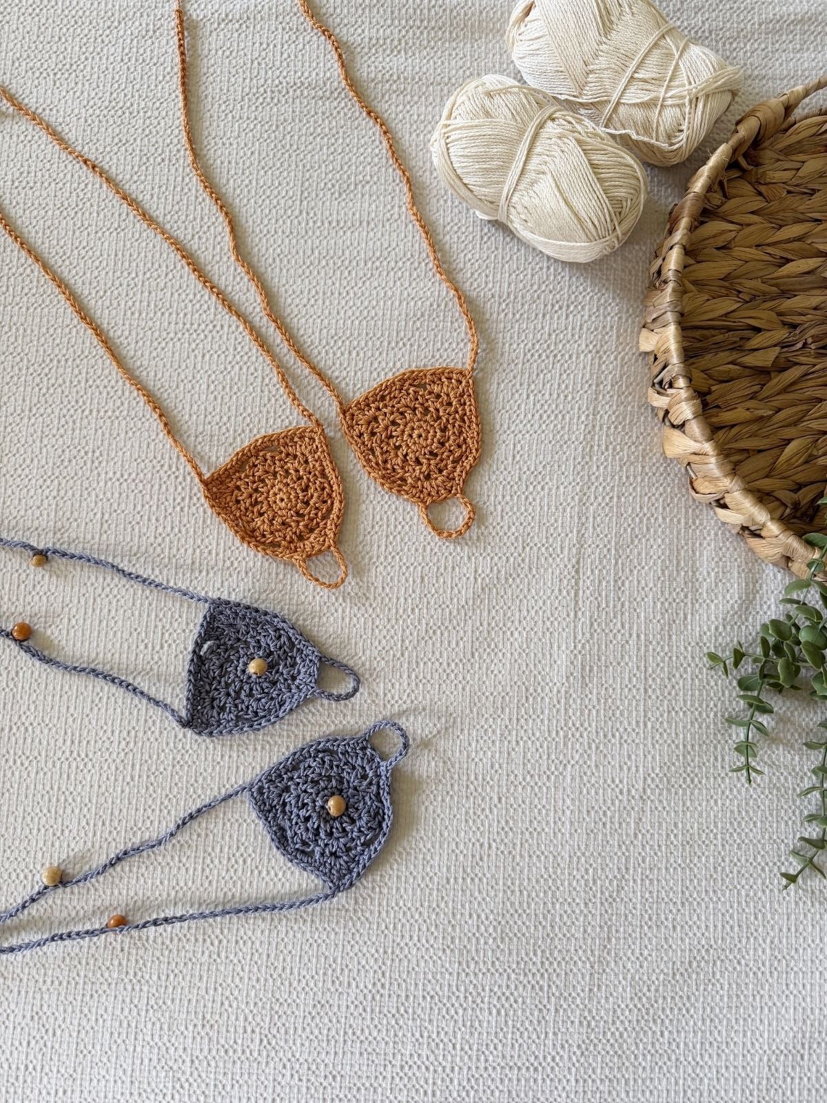 Two pairs of crocheted triangle-shaped barefoot sandals with beads, two balls of beige yarn, and a woven basket rest on a textured white surface—perfect for trying out a free barefoot crochet sandals pattern.