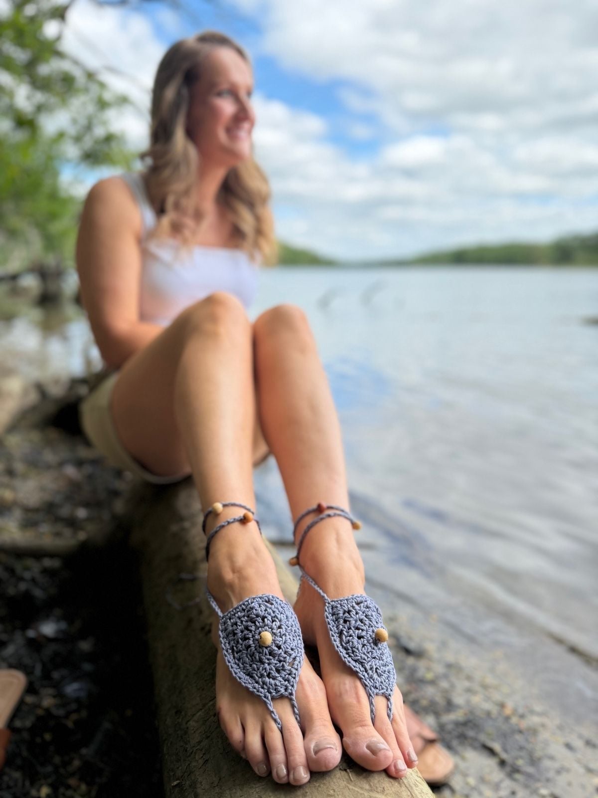 A woman sits on a log by a lake, wearing a white tank top, shorts, and crocheted barefoot sandals with beads made from a free barefoot crochet sandals pattern. The background shows water, trees, and a cloudy sky.