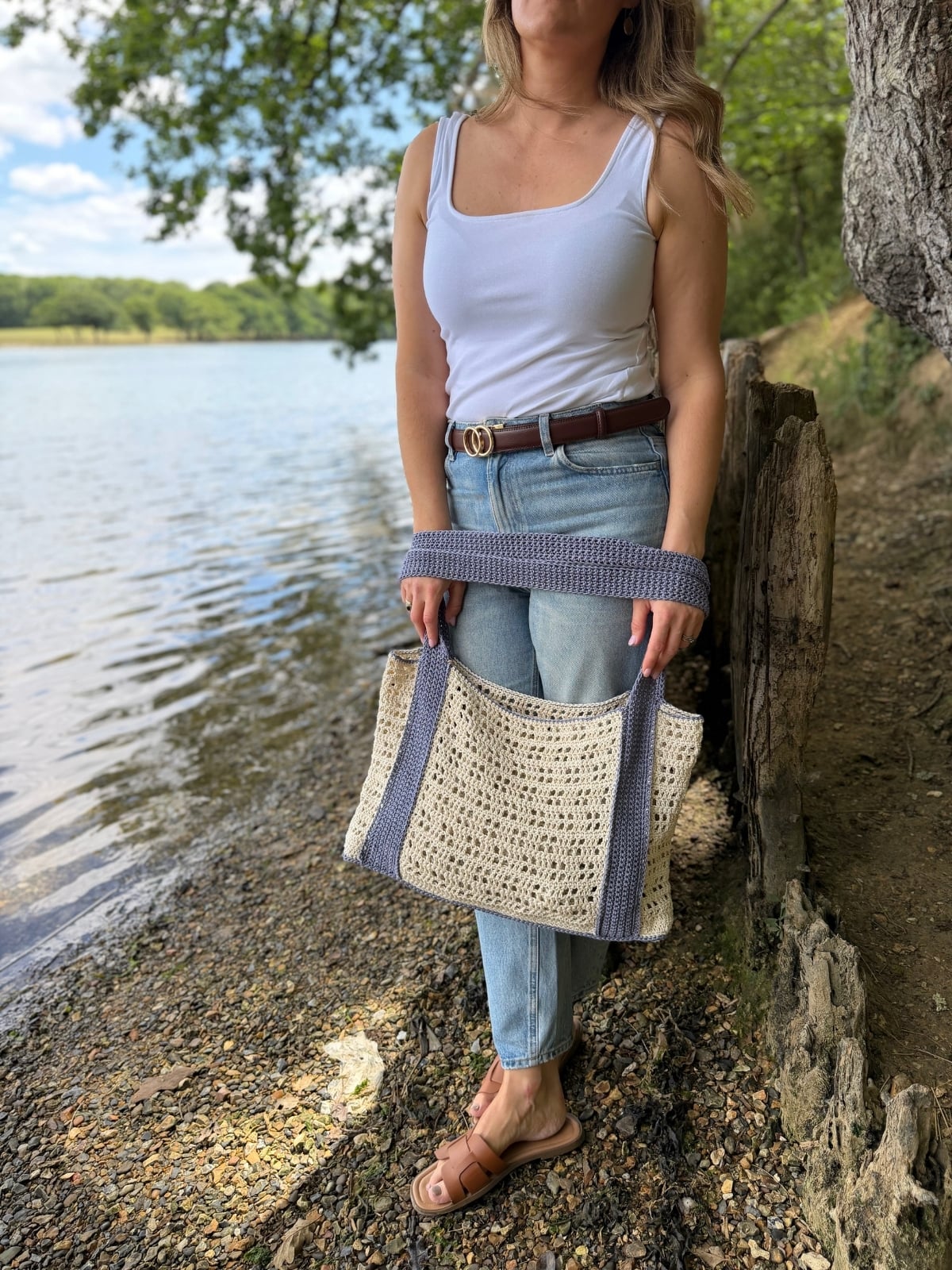 A woman in a white tank top, jeans, and sandals stands by a lake holding a woven bag with blue straps, inspired by a free crochet beach bag pattern.
