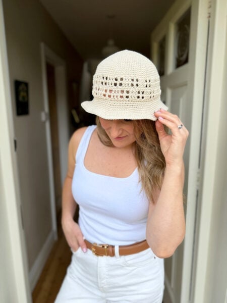 Woman wearing a white tank top, white pants, and a brown belt, adjusting a beige hat made from a Free Crochet Sun Hat Pattern while standing in a hallway.