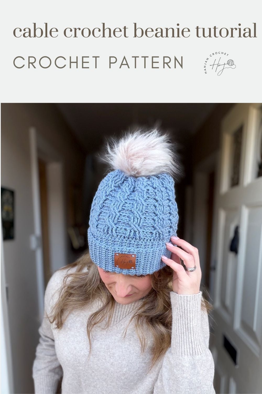 Person wearing a blue cable crochet beanie with a faux fur pompom, standing indoors. Text above reads "cable crochet beanie tutorial" and "free crochet cable hat pattern.