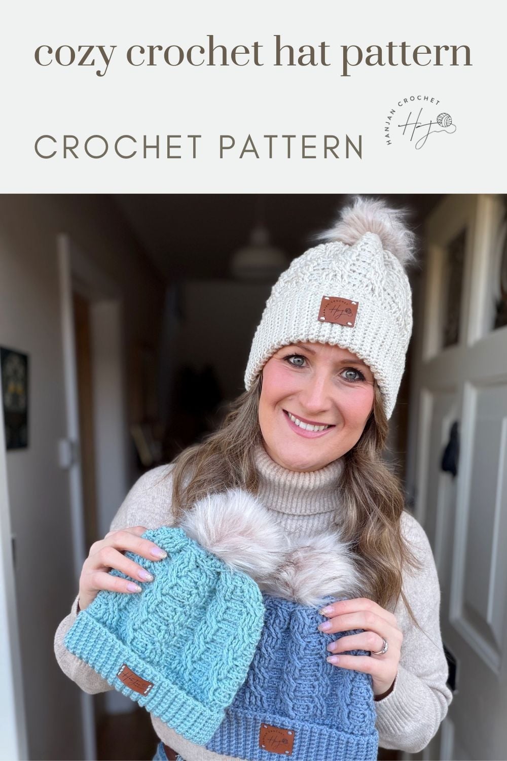 Woman in a beige turtleneck holds two crocheted hats, one blue and one teal, while wearing a white cable hat with a pom-pom, sharing her cozy and free crochet cable hat pattern.
