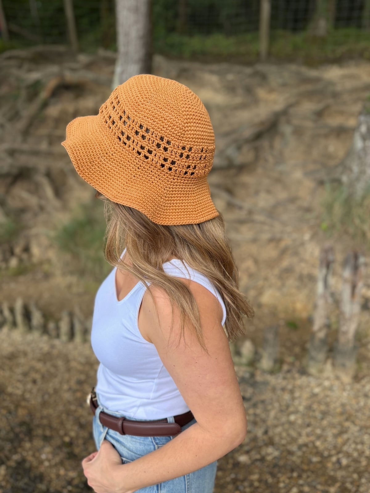 A person wearing a light blue sleeveless top, jeans, and an orange crochet hat—made from a Free Crochet Sun Hat Pattern—stands outdoors on a dirt path, facing away from the camera.
