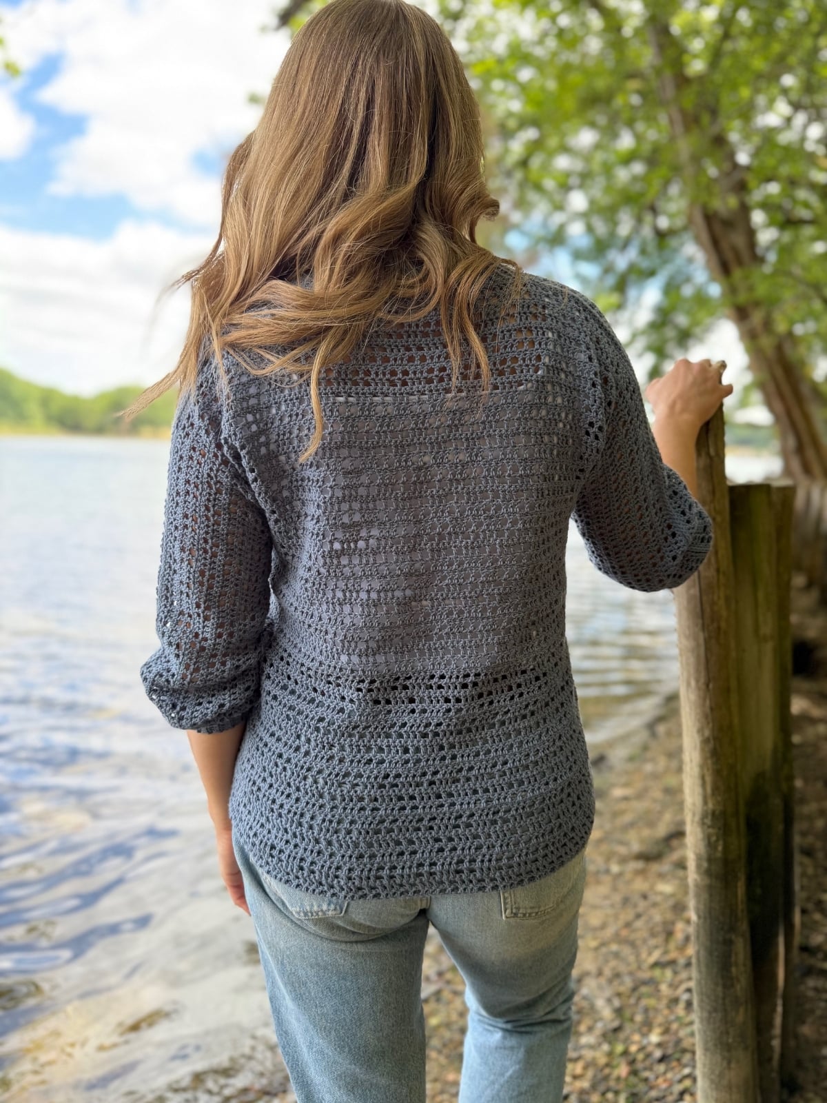 A person with wavy brown hair, wearing a blue summer sweater crochet pattern and jeans, stands by a lakeshore, facing away, holding a wooden post. Trees and water are visible in the background.