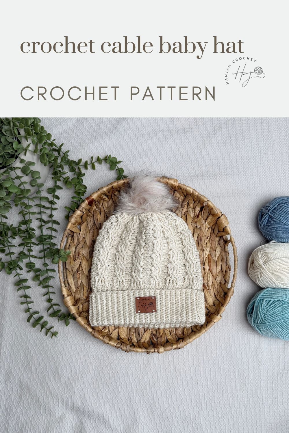 A crocheted baby hat with a cable pattern and faux fur pom-pom sits in a woven basket, next to blue and beige yarn balls—perfect inspiration for your next project using a free crochet cable hat pattern.