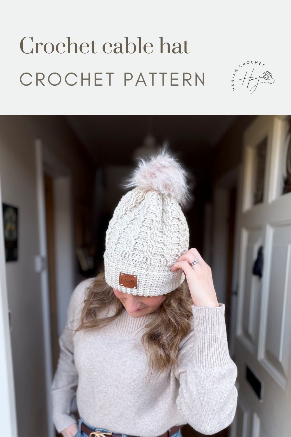 A person wears a cream-colored crochet cable hat with a faux fur pompom, standing indoors. The image highlights a free crochet cable hat pattern perfect for cozy and stylish handmade accessories.