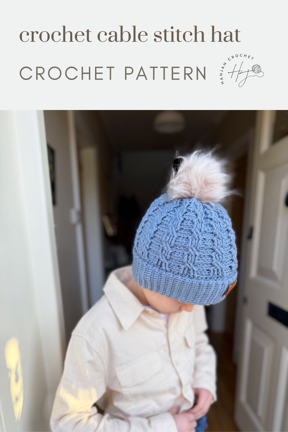 A child wearing a light blue crochet cable stitch hat with a faux fur pom-pom stands indoors, looking down. Text above reads "crochet cable stitch hat FREE CROCHET CABLE HAT PATTERN.