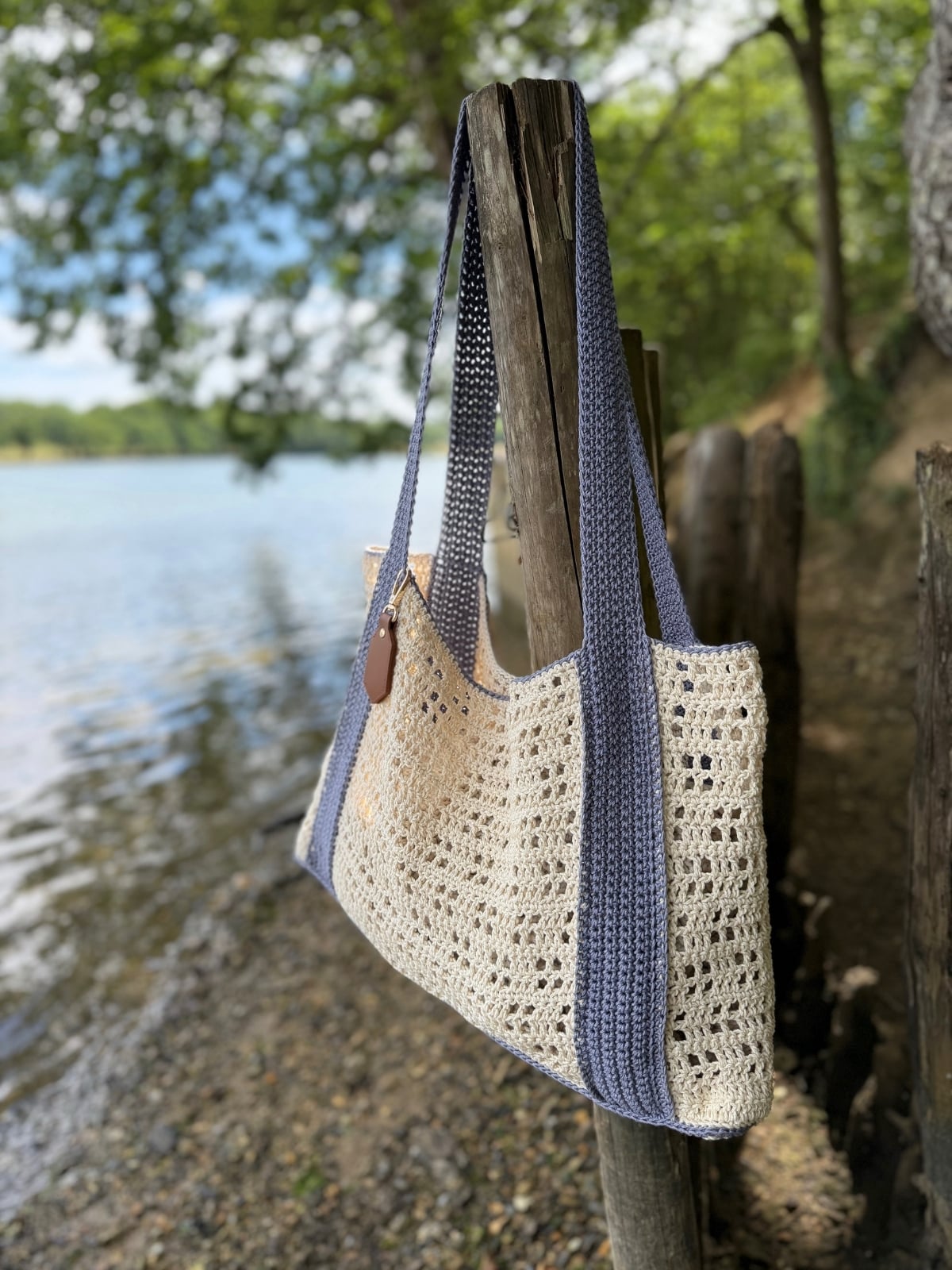 A crocheted beige and blue tote bag, made using a free crochet beach bag pattern, hangs on a wooden post beside a calm lake, with trees and a pebbly shore in the background.