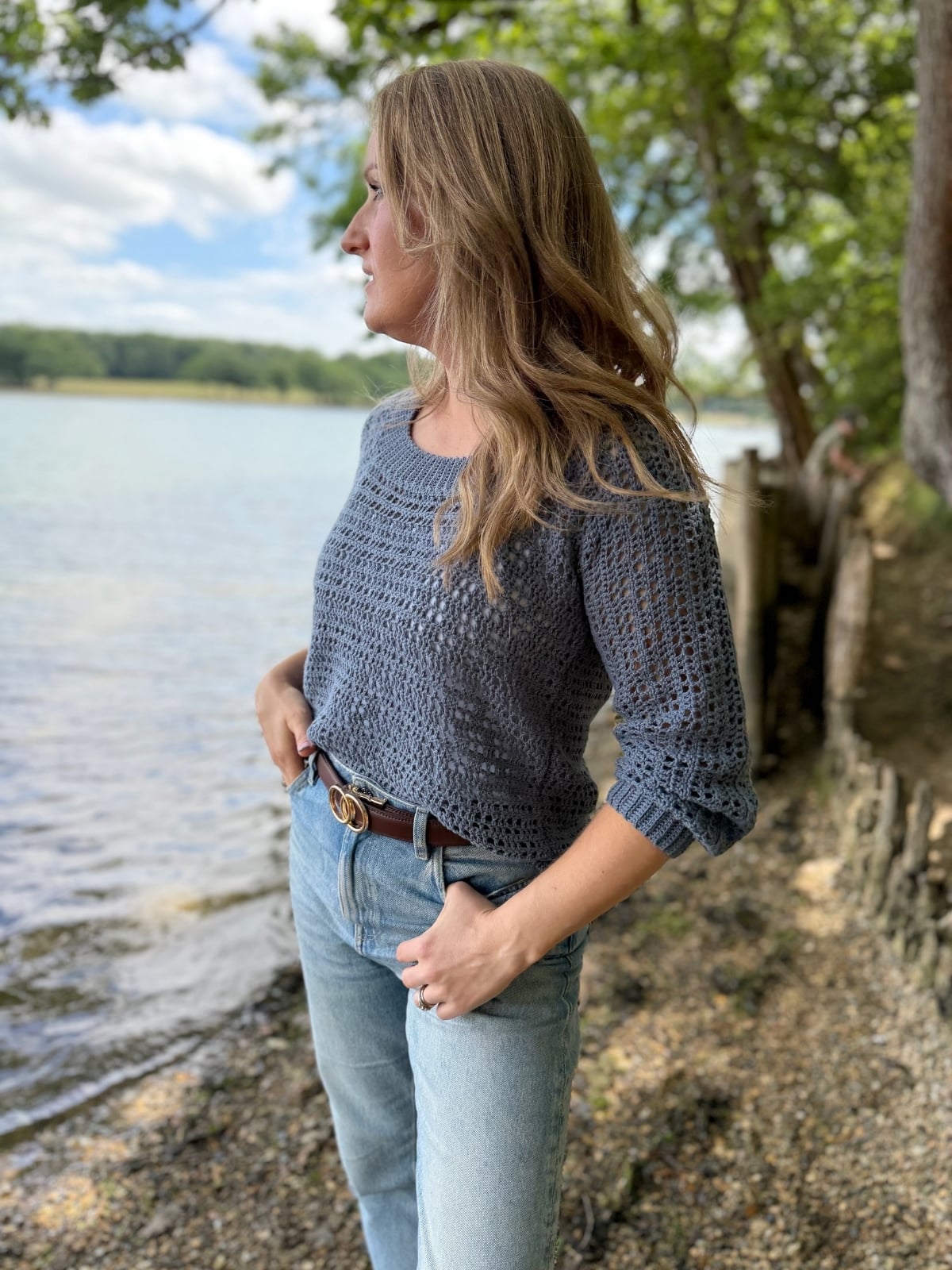 A person in a gray summer sweater crochet pattern and jeans stands by a lake, looking into the distance with hands in pockets. Trees and water are visible in the background.
