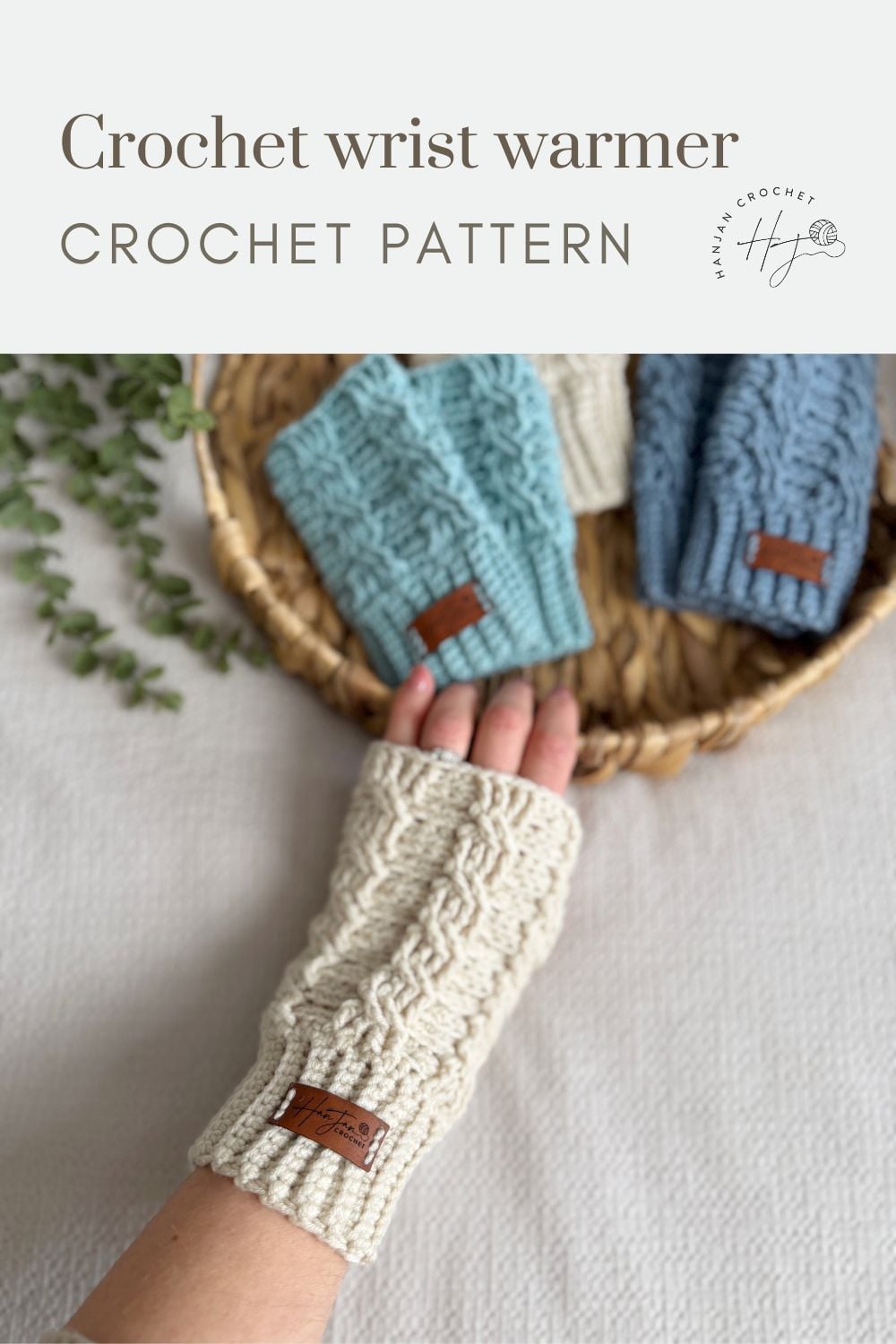 A hand wearing a cream-colored crochet wrist warmer with a brown label rests beside a basket of blue and cream wrist warmers on a white surface—perfect inspiration if you’re looking for a crochet wrist warmers free pattern.
