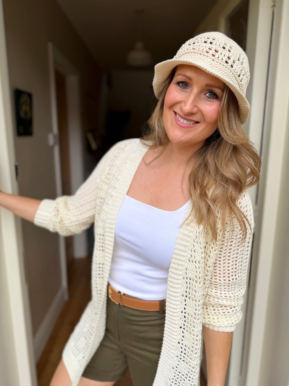 A woman in a cream crochet hat and summer cardigan, white tank top, and olive shorts stands in a doorway, smiling at the camera, showcasing a stylish crochet pattern.