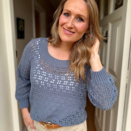 Woman standing indoors, smiling at the camera, with one hand touching her hair. She is wearing a blue crochet sweater inspired by a summer sweater crochet pattern and beige pants.