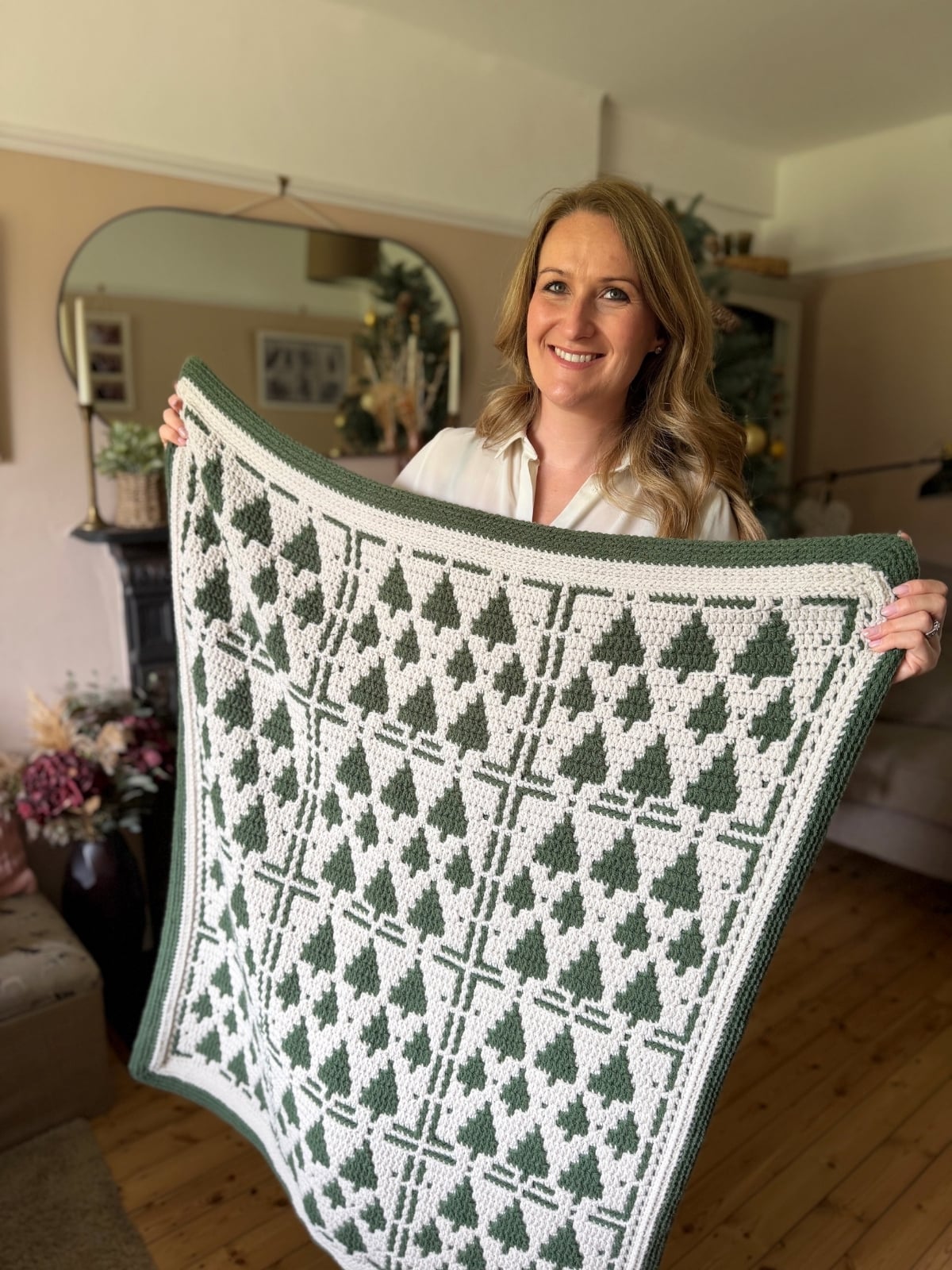 A woman stands indoors holding up a handmade green and white Christmas crochet afghan featuring a festive tree pattern.