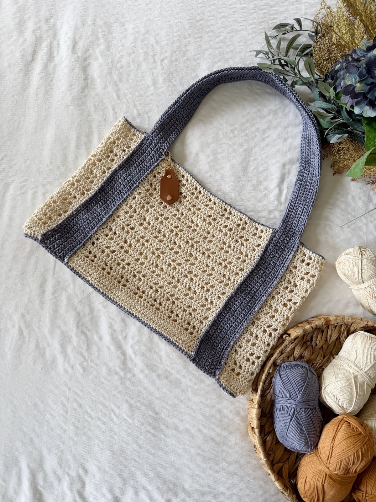 A crochet tote bag made with beige and gray yarn rests on a white surface near a basket of assorted yarn balls and green foliage, inspired by a free crochet beach bag pattern.