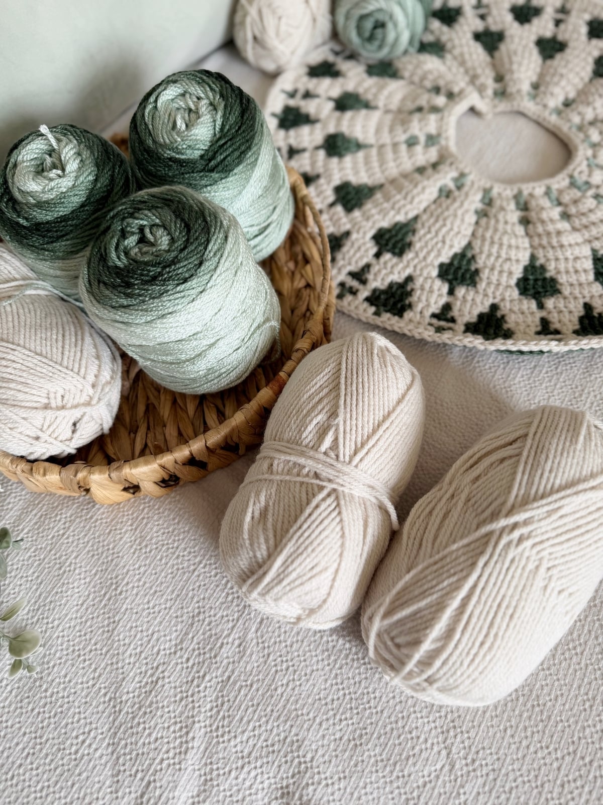 A basket with green and cream yarn balls sits on a textured surface next to two cream yarn skeins, a partially completed crochet project, and a crochet tree skirt pattern.