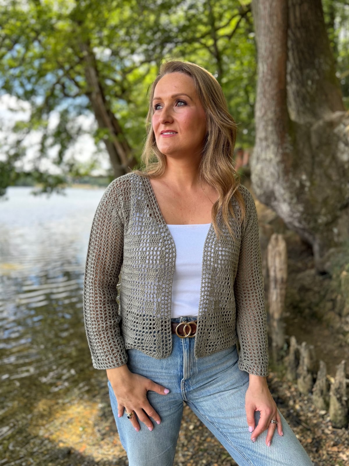 A woman stands near a lakeside, wearing a light gray open-knit summer cardigan crochet pattern over a white top and blue jeans, with trees and water in the background.