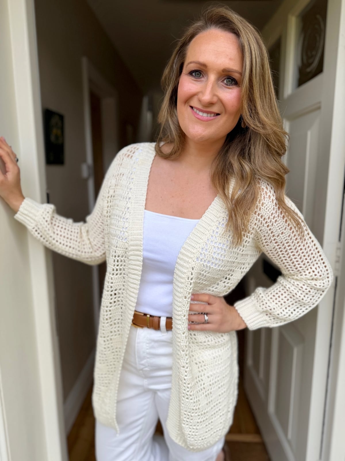 A woman with long blonde hair, wearing a white crochet cardigan inspired by a summer cardigan crochet pattern over a white top and white pants, stands in a hallway and smiles at the camera.
