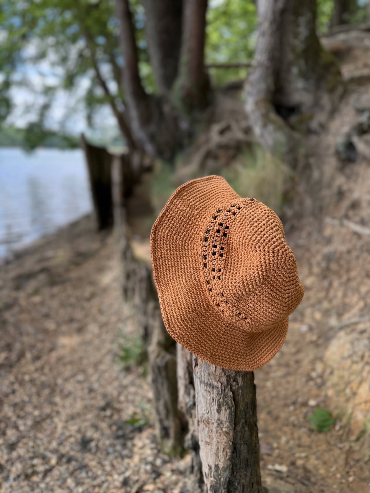 A brown woven hat, reminiscent of a Free Crochet Sun Hat Pattern, hangs on a wooden post along a pebbly lakeshore, with trees and water visible in the background.