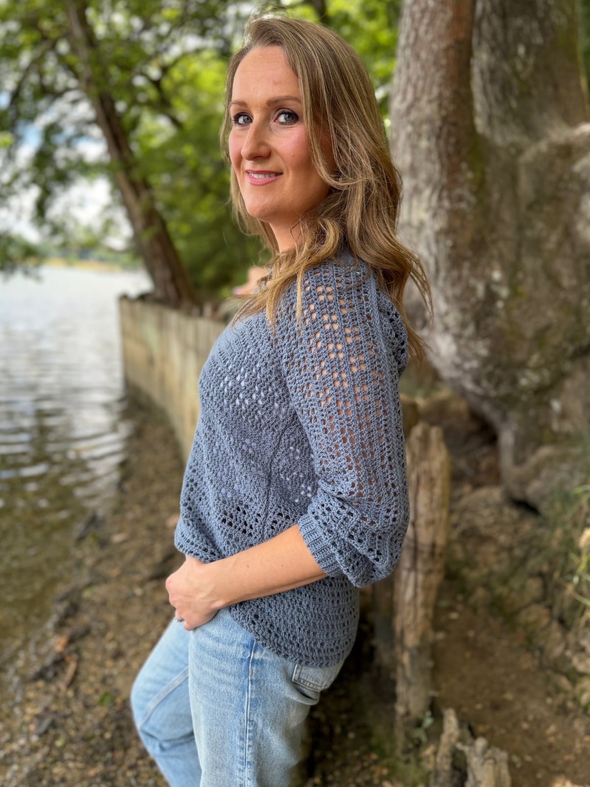 A woman with long, wavy hair stands by a lakeside, wearing a blue summer sweater crochet pattern and jeans, with trees and water in the background.