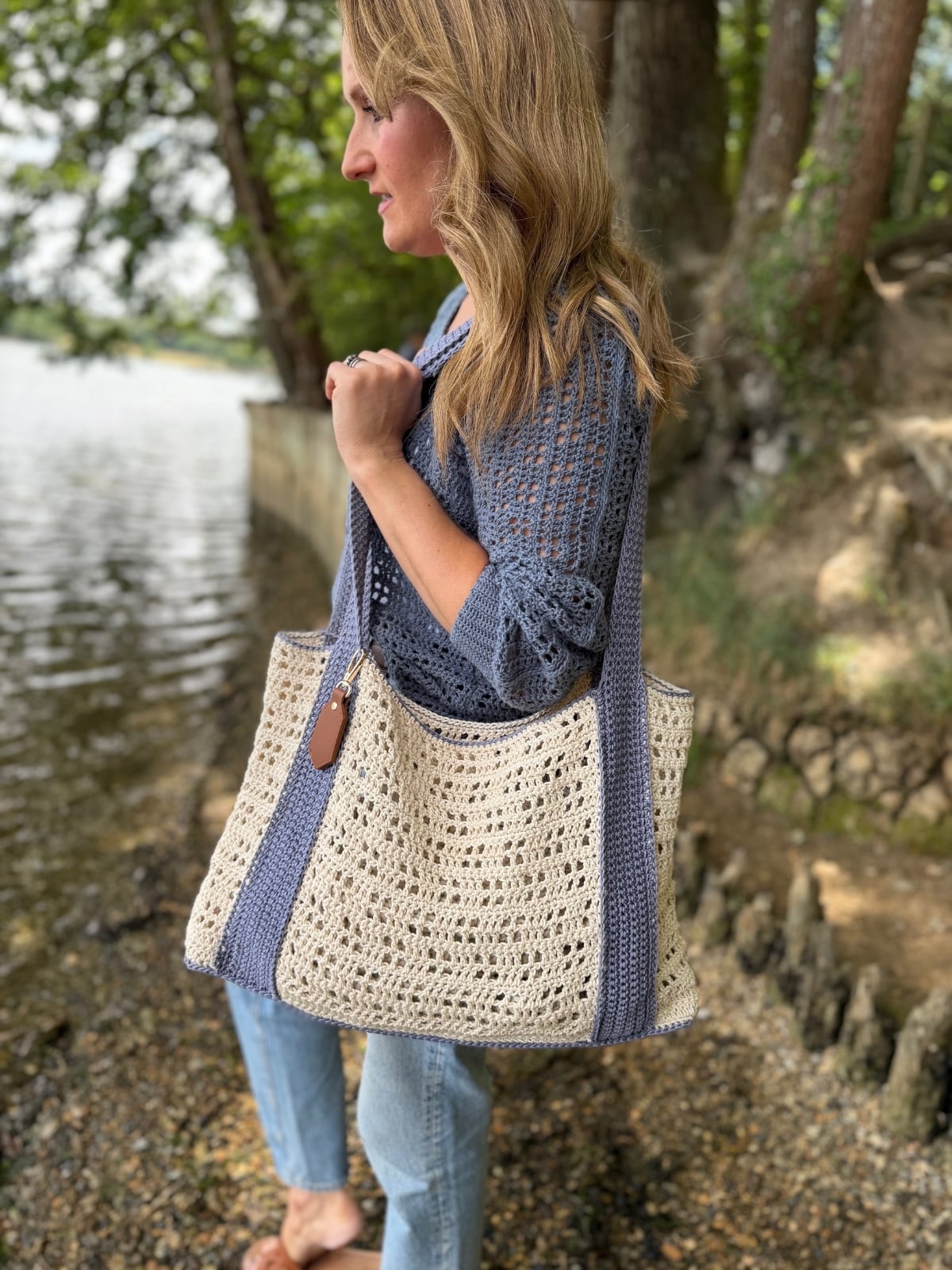 A woman stands by the water on a pebbled shore, wearing a blue sweater, jeans, and carrying a large tote made from a free crochet beach bag pattern in blue and cream colors.